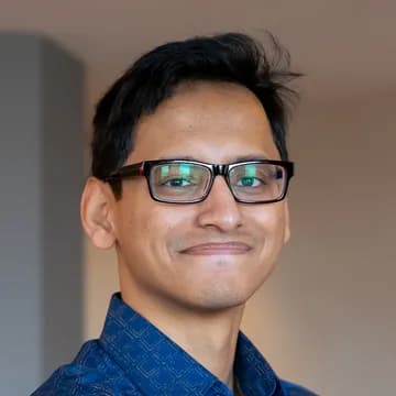 Abir, a brown man with black short hair is smiling at the camera. He wears glasses and a blue shirt.