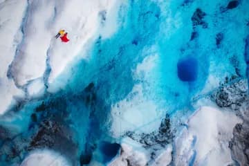 Drone shot of a man in a yellow jacket and a woman in a red dress lying on glacial ice next to a blue glacial water body.