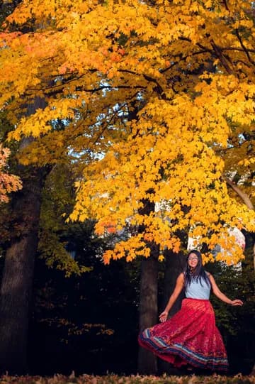 A woman in a red skirt and white top standing next to a series of trees with yellow and orange leaves, and with bright red fallen leaves covering the ground in front of her.