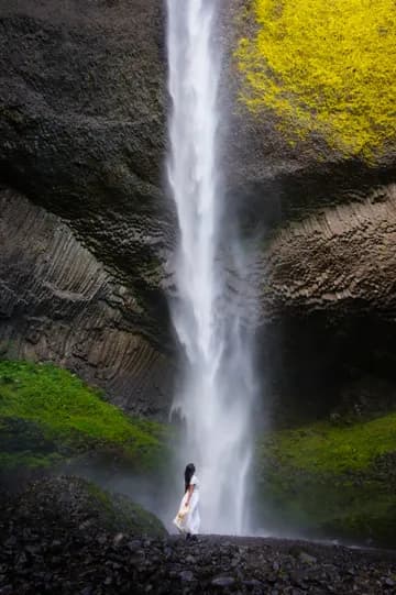 A woman wearing a white dress and straw hat in front of horsetail-shaped waterfall.