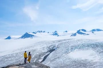 A man wearing a yellow jacket and a woman wearing white top and yellow skirt standing in the distance looking towards a vast ice field.