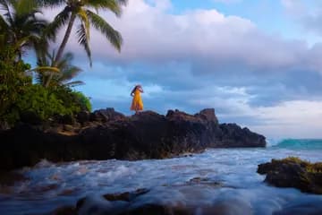 A woman in yellow dress standing on a rocky beach looking towards the ocean waves, with a big wave just crashing over the rock.