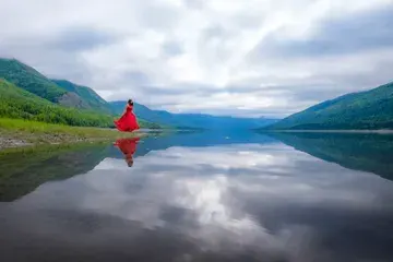 A woman in a red dress looking towards a lake. The pristine lake water has a perfect mirror-like reflection of the woman, the sky and nearby hills.