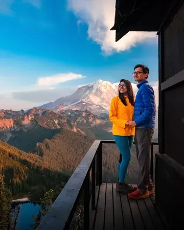 A woman in yellow jacket and a man in blue jacket smilingly holding hands. Snow-covered Mount Rainier can be seen in the background.