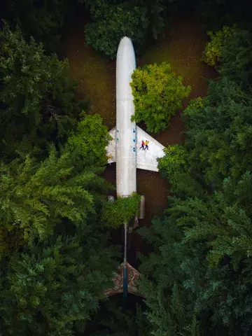Drone shot of an aeroplane surrounded by dense forest, with a man and a woman lying on the right wing.