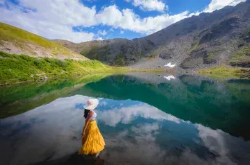 A woman in an yellow skirt looking towards a lake. The pristine blue lake water has a perfect reflection of the blue sky and nearby hills.