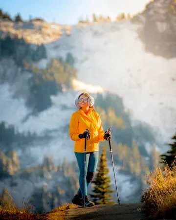 A woman in a yellow jacket smilingly holding her hiking poles, with warm afternoon light falling on her face and a snow-covered mountain in the backdrop.