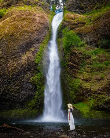 A woman wearing a white dress with a straw hat in her right hand in front of long waterfall