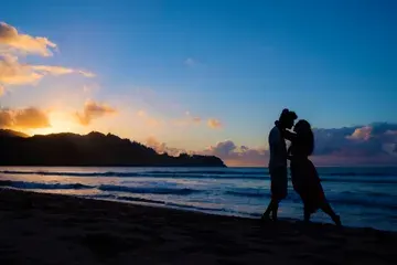 Silhouettes of a man and a woman about to kiss. Pacific Ocean in the backdrop. The sky is deep blue and last rays of sunset bleeding through from behind mountains on the left.